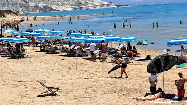 En strand med mennesker, parasoller og en klippe i baggrunden.