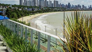Ein Strand mit Stadtansicht im Hintergrund, ein Balkon mit Pflanzen und ein Poolbereich.