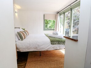 A bedroom with a bed, pillows, a green throw blanket, a window with curtains, and a view of greenery outside. - Vintner's Cottage (Norwich)