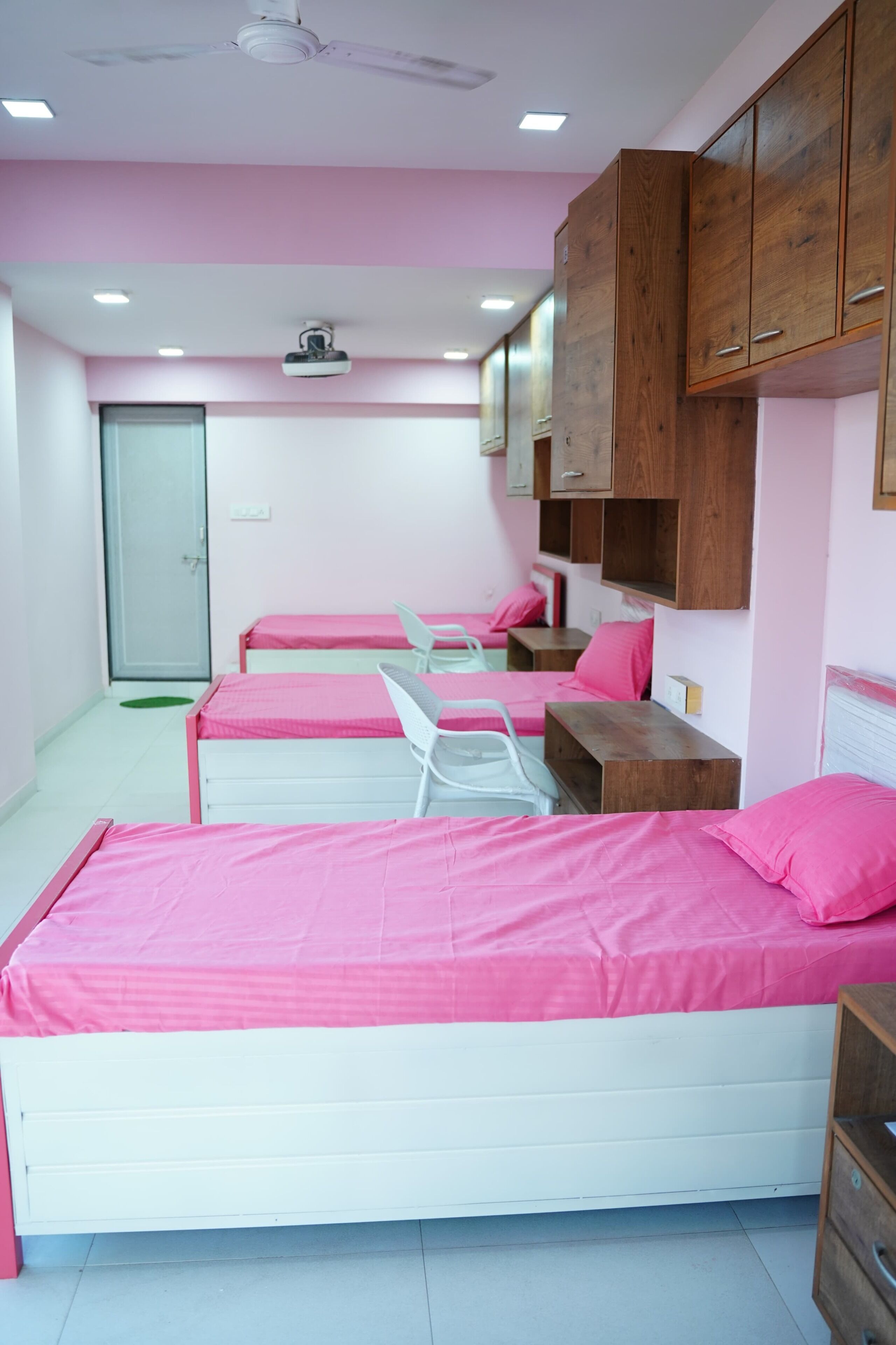 A room with three beds, each with a pink mattress and white frame, wooden cabinets above, and a ceiling fan.