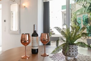 Een tafel met een wijnfles en twee glaasjes, een kamerplant en een raam met uitzicht op groen.