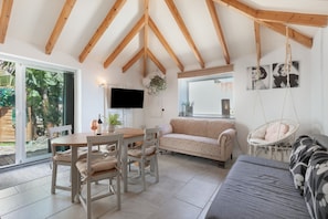 Een ruime kamer met een houten plafond, een eettafel met stoelen, een bank, een hangstoel en een aan de muur bevestigde televisie.