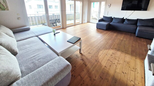 Een ruime woonkamer met een bank, een salontafel en een aan de muur bevestigde televisie.