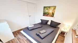 Een moderne slaapkamer met een bed, kussens, een nachtkastje met een lamp, een kleine plant en een schilderij aan de muur.