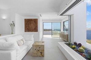 A modern living room with a white sofa, a decorative rug, and a view of the sea through large windows. - Infinity House, Perched majestically on a cliff along the island’s western coast (Paros)
