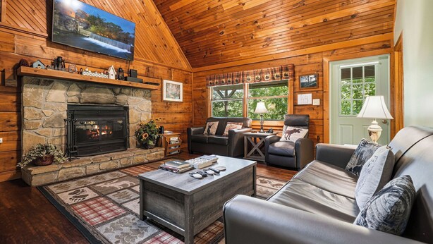 A cozy log cabin interior with a stone fireplace, leather sofas, a flat-screen TV, and a large window with a view of greenery. - Cozy* Secluded*Firepit*Hot Tub*Seeps 7 (Pigeon Forge)