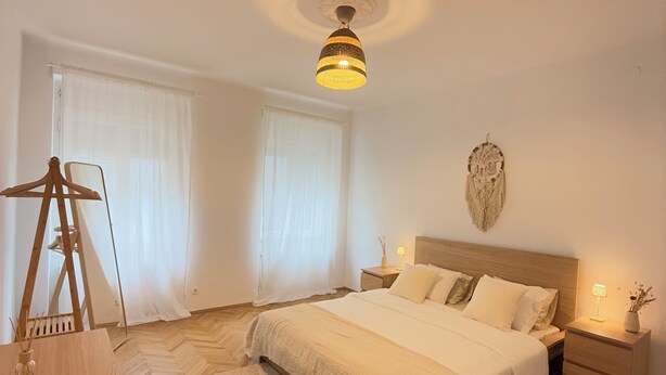 A bedroom with a bed, bedside tables, a hanging light fixture, a wall decoration, and a wooden dresser. - Stylish apartment in the city center (Graz)