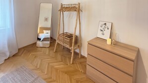 A room with a wooden herringbone floor, a wooden dresser, a wicker chair, a bed with a white headboard, and a wall-mounted mirror. - Stylish apartment in the city center (Graz)
