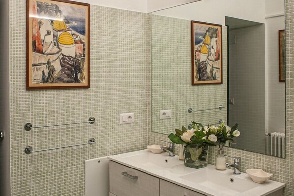 A bathroom with a tiled wall, a sink with a mirror, and two framed artworks.