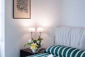 A neatly arranged bedroom with a bed, two bedside lamps, a vase with flowers, and a framed artwork on the wall.