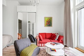 A room with a bed, a gray armchair, a red sofa, a white coffee table with books, a painting, and a large window with curtains. - Balcony Studio | Rooftop Views | Tram 200m | Lift (Helsinki)