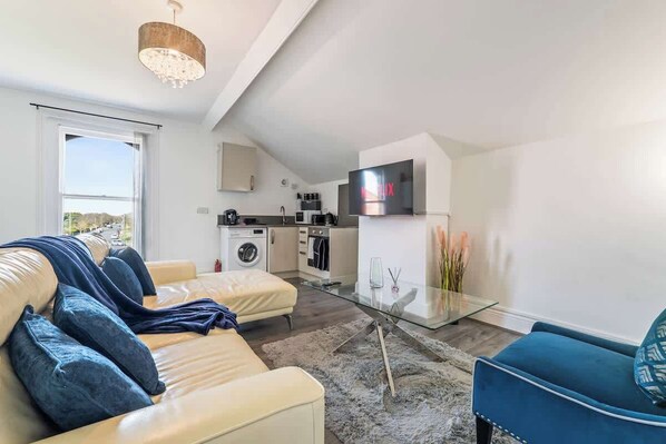 Un salon moderne avec un canapé, une table basse en verre et un fauteuil bleu. On aperçoit une cuisine équipée d’un lave-linge et d’un téléviseur au fond.