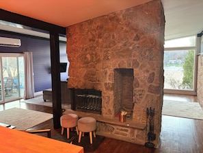 Une cheminée en pierre avec un feu, deux tabourets et un téléviseur dans un salon moderne doté de grandes fenêtres.