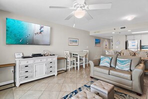 Un vaste espace de vie agrémenté d’un ventilateur de plafond, d’une grande œuvre d’art sur le thème de la plage, d’un coin repas avec des chaises blanches et d’un canapé beige orné de coussins à motifs.
