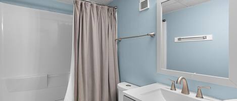 Un baño con cortina de ducha, inodoro, lavamanos con espejo y toallero.
