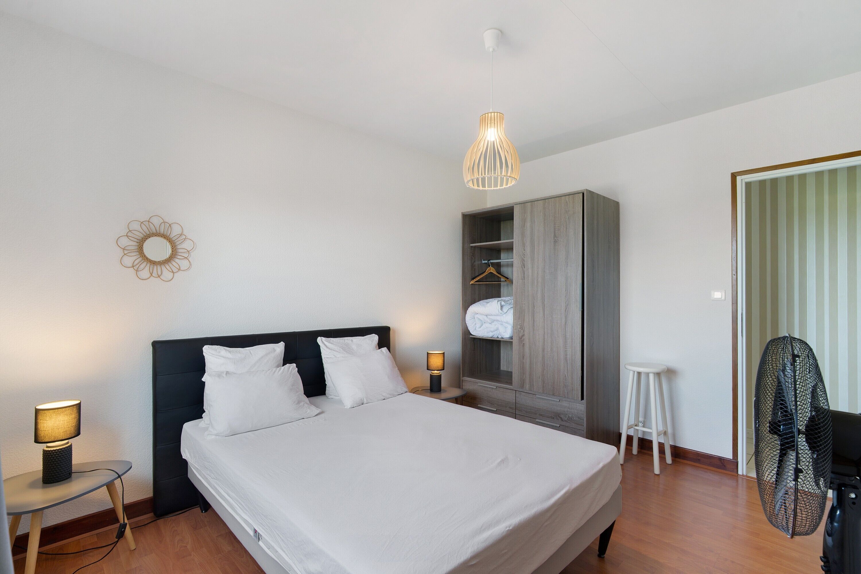 A neatly arranged hotel room with a bed, bedside lamps, a wardrobe, a small stool, and a fan.