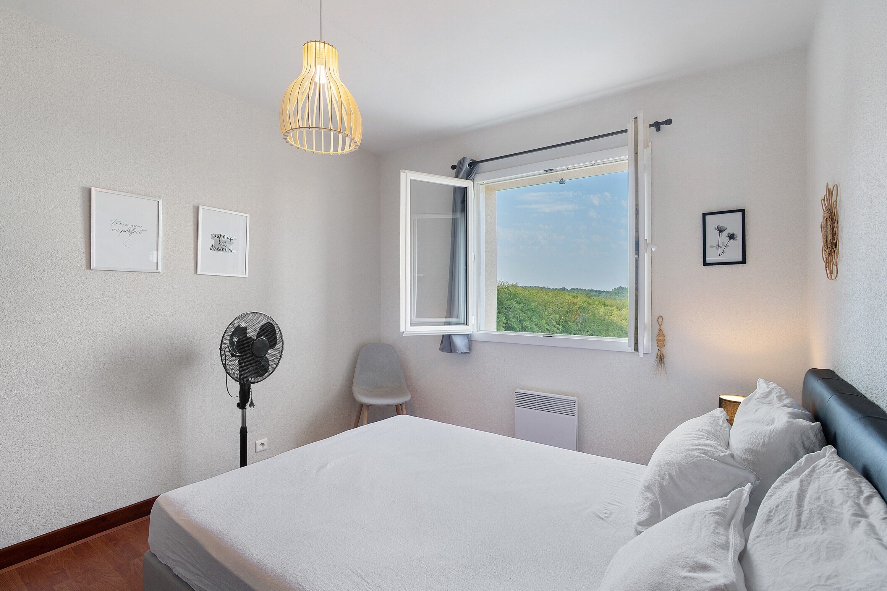 A bedroom with a bed, a standing fan, a window with an open shutter, and wall-mounted artwork.