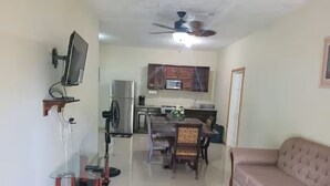 Una sala de estar con sofá, televisor montado en la pared, ventilador de techo y una zona de cocina con armarios y electrodomésticos.