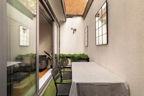 Ein schmaler Außenbereich mit Tisch und Stühlen, eine kleine Gartenanlage und eine Glastür, die zu einem Innenraum führt.
