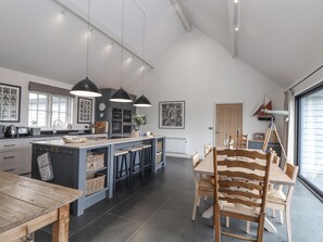 A modern kitchen with a large island, pendant lights, and a dining area with wooden chairs. - Wood Meadow (Helston)