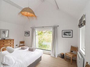 A bedroom with a bed, a hanging light, a window with curtains, a chair, a radiator, and framed pictures on the wall. - Wood Meadow (Helston)