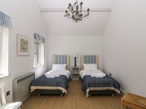 A bedroom with two beds, a radiator, a chandelier, a window with curtains, and a framed picture on the wall. - Wood Meadow (Helston)