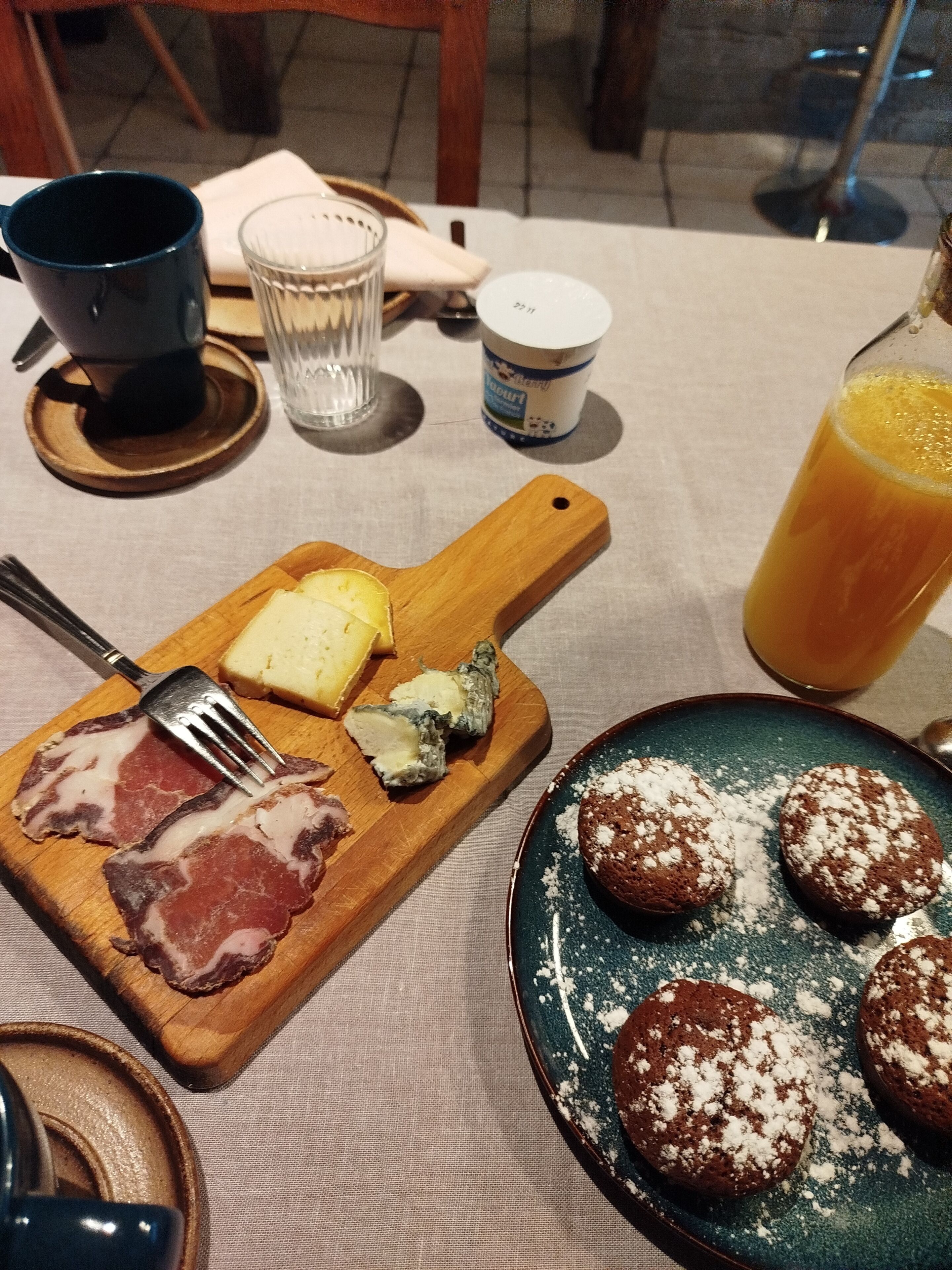 Mesa de café da manhã com tábua de queijos e presunto, prato de biscoitos com açúcar e uma taça de suco de laranja, além de uma xícara de café.