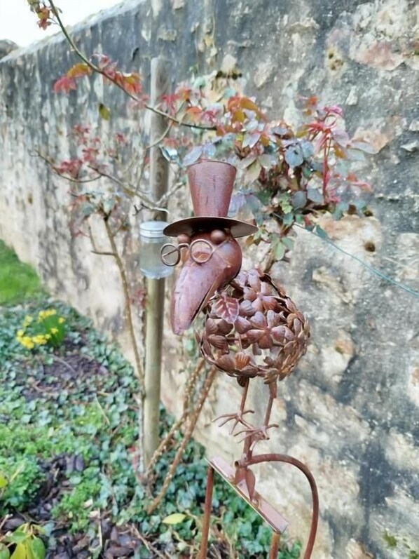 A metal sculpture of a bird with a vase on its head, set against a wall with ivy and a garden. - Château Besson (Désertines)