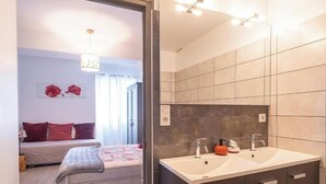 Een slaapkamer met een bed, een nachtkastje en een wandlampje. Er is een raam met gordijnen, een vastzittende lamp en een lijst met een afbeelding. De badkamer heeft een grote spiegel, een wastafel en een betegelde muur.
