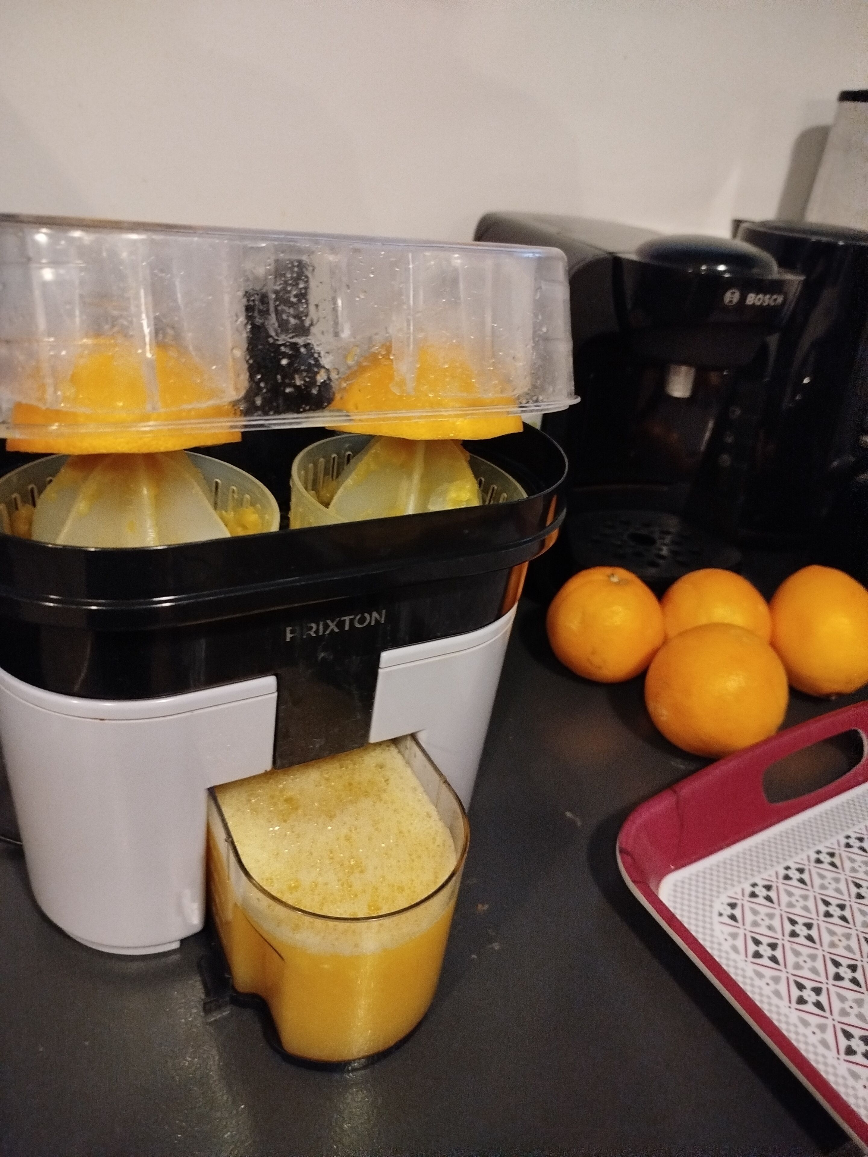 A kitchen with a juicer, a glass of juice, and several oranges.