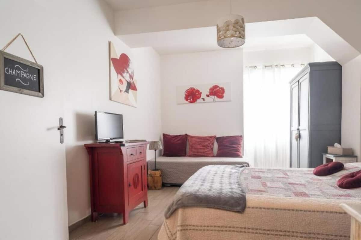 A bedroom with a bed, a TV, a red dresser, a chandelier, and a door leading to another room.