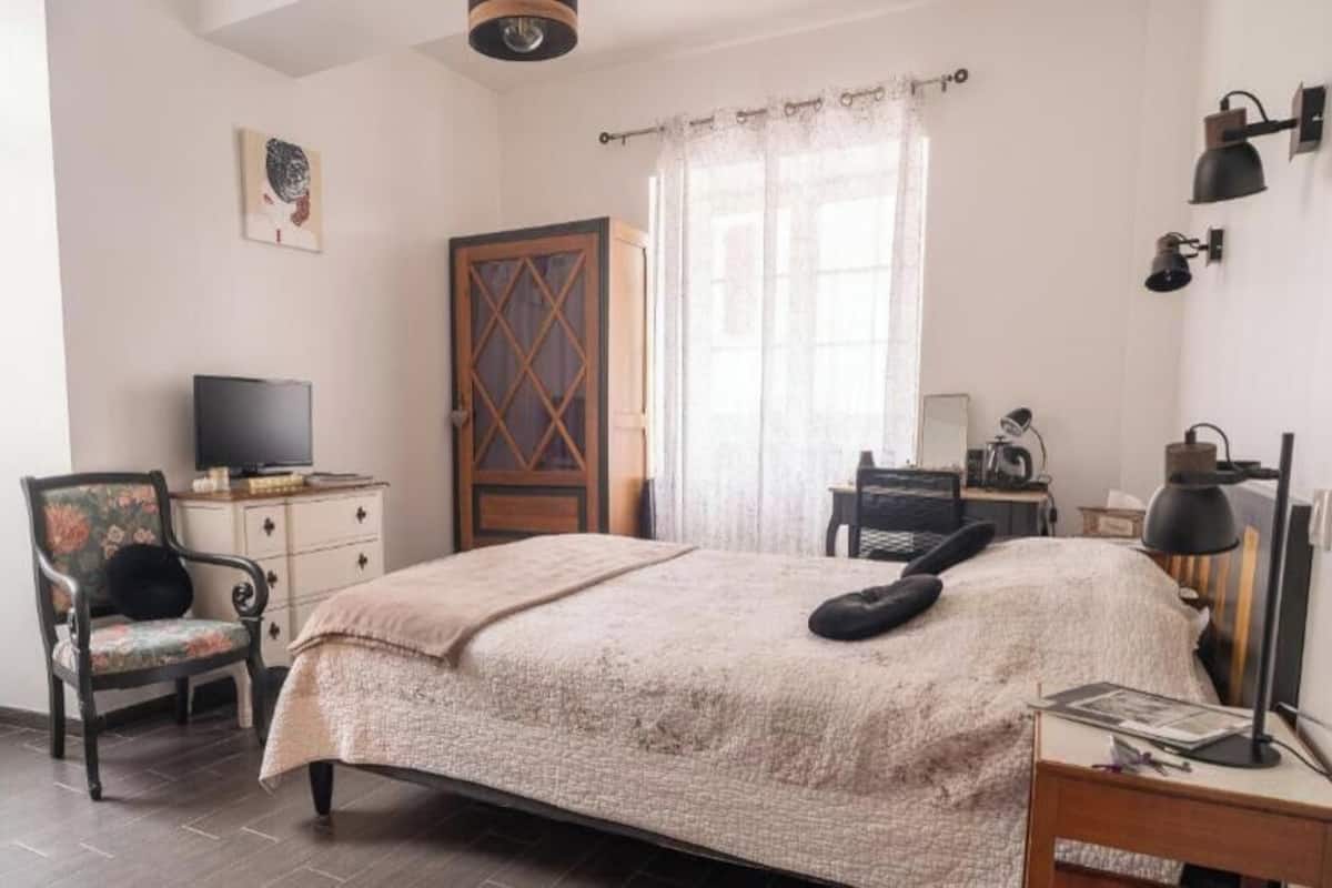 A bedroom with a bed, a chair, a television, a wardrobe, and a desk with a lamp.