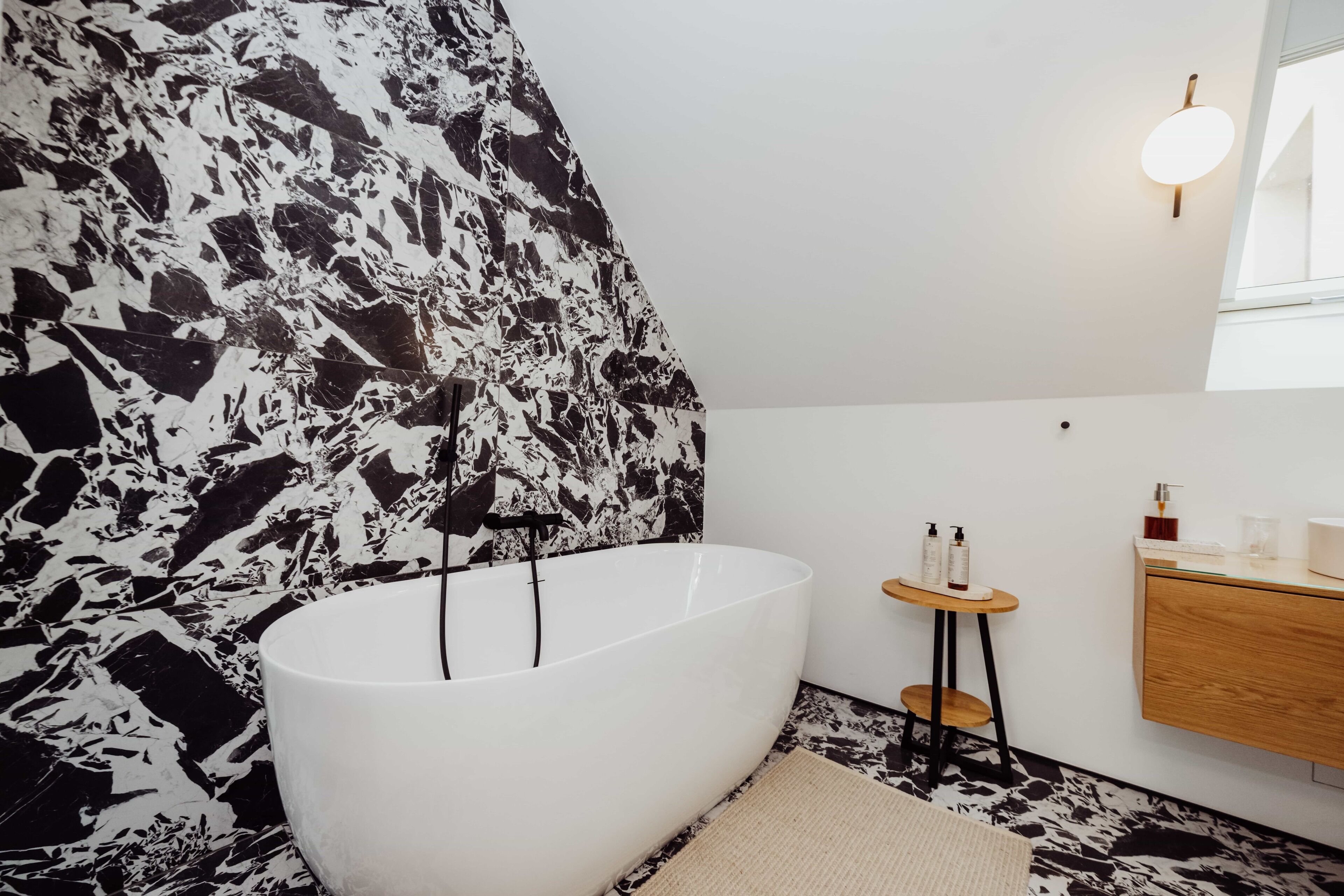Un baño moderno con una bañera independiente, un tocador de madera pequeño y una pared con diseño de mármol.