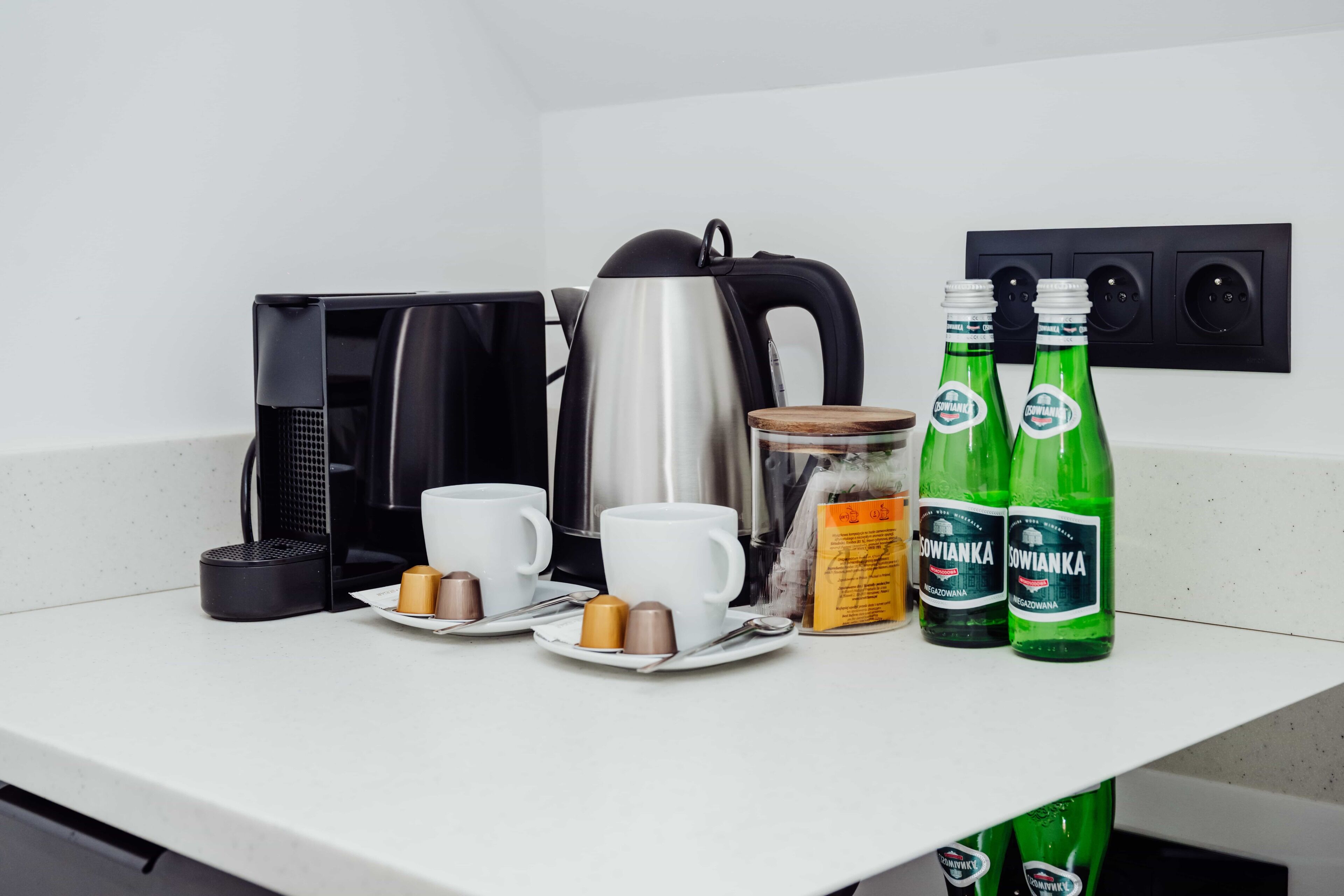 A kitchen counter with a coffee maker, two cups, two bottles of Sowianka, a jar, and a small container.