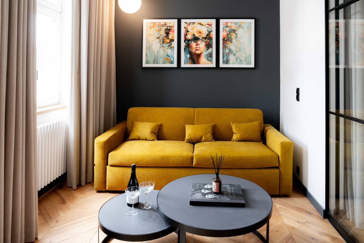 Ein Wohnzimmer mit einem senfgelben Sofa, zwei runden Tischen und drei gerahmten Kunstwerken an der Wand.