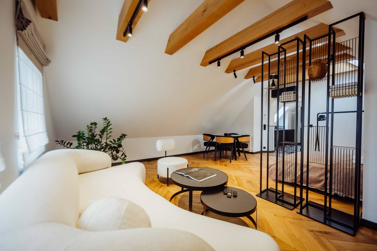 Ein modernes Loft-Zimmer mit hoher Decke, freiliegenden Holzbalken, einem großen weißen Sofa, einem runden Tisch und einer schwarzen Metallabtrennung.