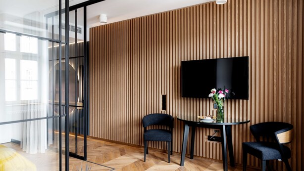 A modern room with a large TV mounted on a wooden wall, two chairs, a table with a vase, and a glass partition. - Grodzka Apartments (Jelenia Góra)
