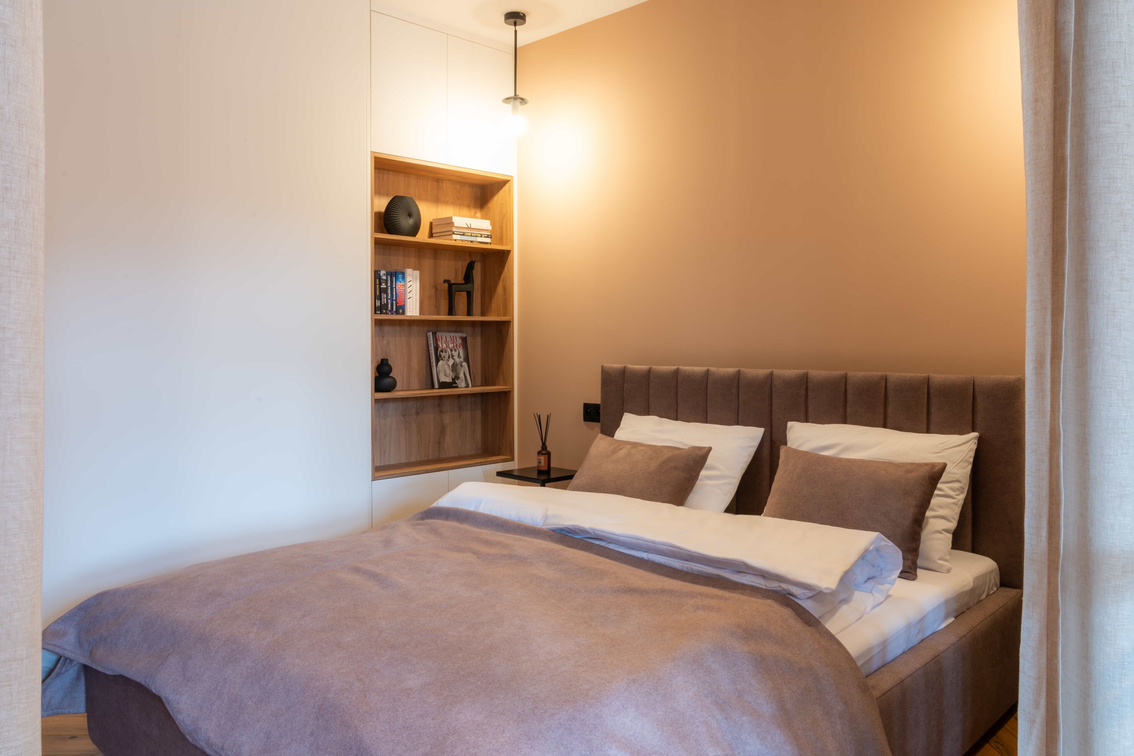 A modern bedroom with a bed, a nightstand, a shelf with books and decorative items, and a wall-mounted light.