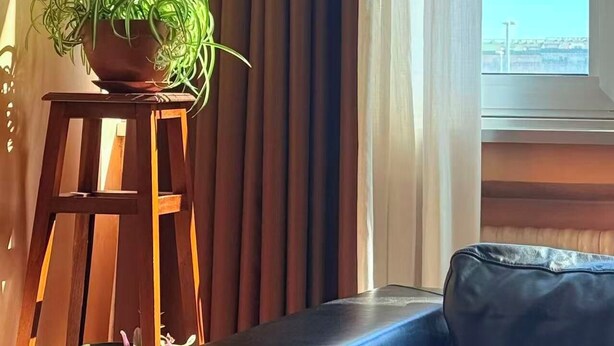 Quarto com janela, cortinas, um sofá, uma pequena mesa com uma planta e uma tigela decorativa.