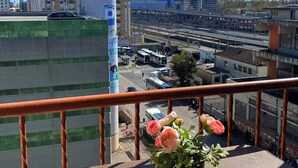 Uma varanda com um vaso de flores, vista de uma paisagem urbana com edifícios e guindastes, e um céu azul límpido.
