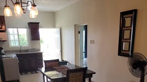 A dining area with a table and chairs, a refrigerator, a kitchen area, and a fan. - Kingsbird By The Sea- Cozy 1 Bedroom (Runaway Bay)