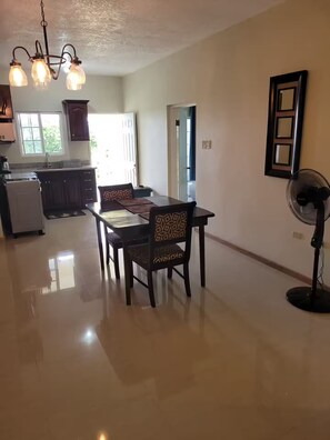 A dining area with a table and chairs, a refrigerator, a kitchen area, and a fan. - Kingsbird By The Sea- Cozy 1 Bedroom (Runaway Bay)