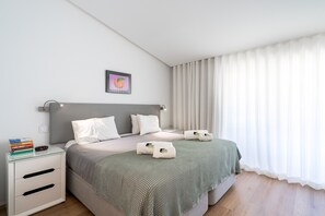 A modern bedroom with a bed, bedside tables, a bedside lamp, a framed picture, and a window with curtains. - Quinta do Lago Charming Apartment (S53) (Quinta do Lago)