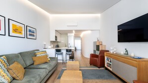 Ein modernes Wohnzimmer mit Sofa, Hocker, Fernseher und einer Küche im Hintergrund.