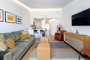 A modern living room with a sofa, ottoman, TV, and a kitchen in the background. - Quinta do Lago Charming Apartment (S53) (Quinta do Lago)