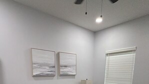 Une chambre à coucher comprenant un lit, deux œuvres d’art accrochées au mur, une lampe de chevet et une fenêtre avec des stores.