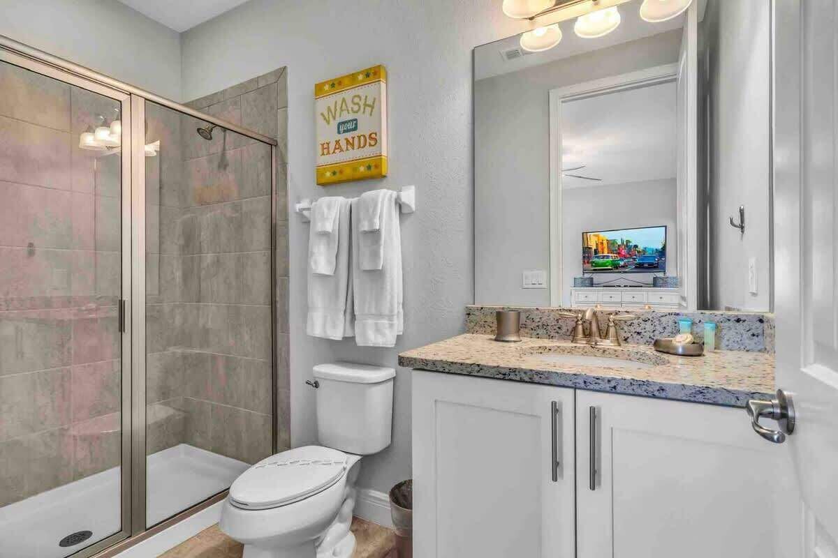 A modern bathroom with a glass shower enclosure, a toilet, a sink with a granite countertop, and a mirror with a 'WASH HANDS' sign.