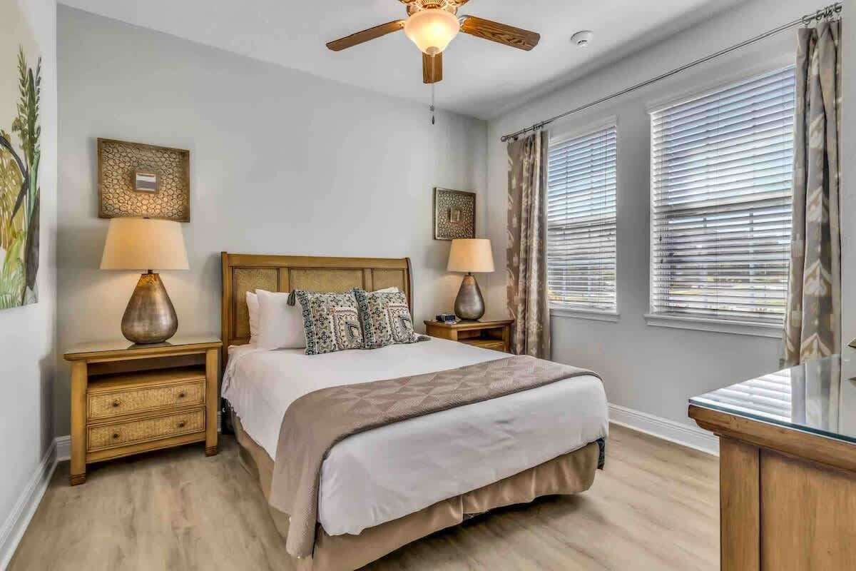 Un dormitorio con cama, dos mesitas de noche con lámparas, un ventilador de techo y una ventana con persianas.
