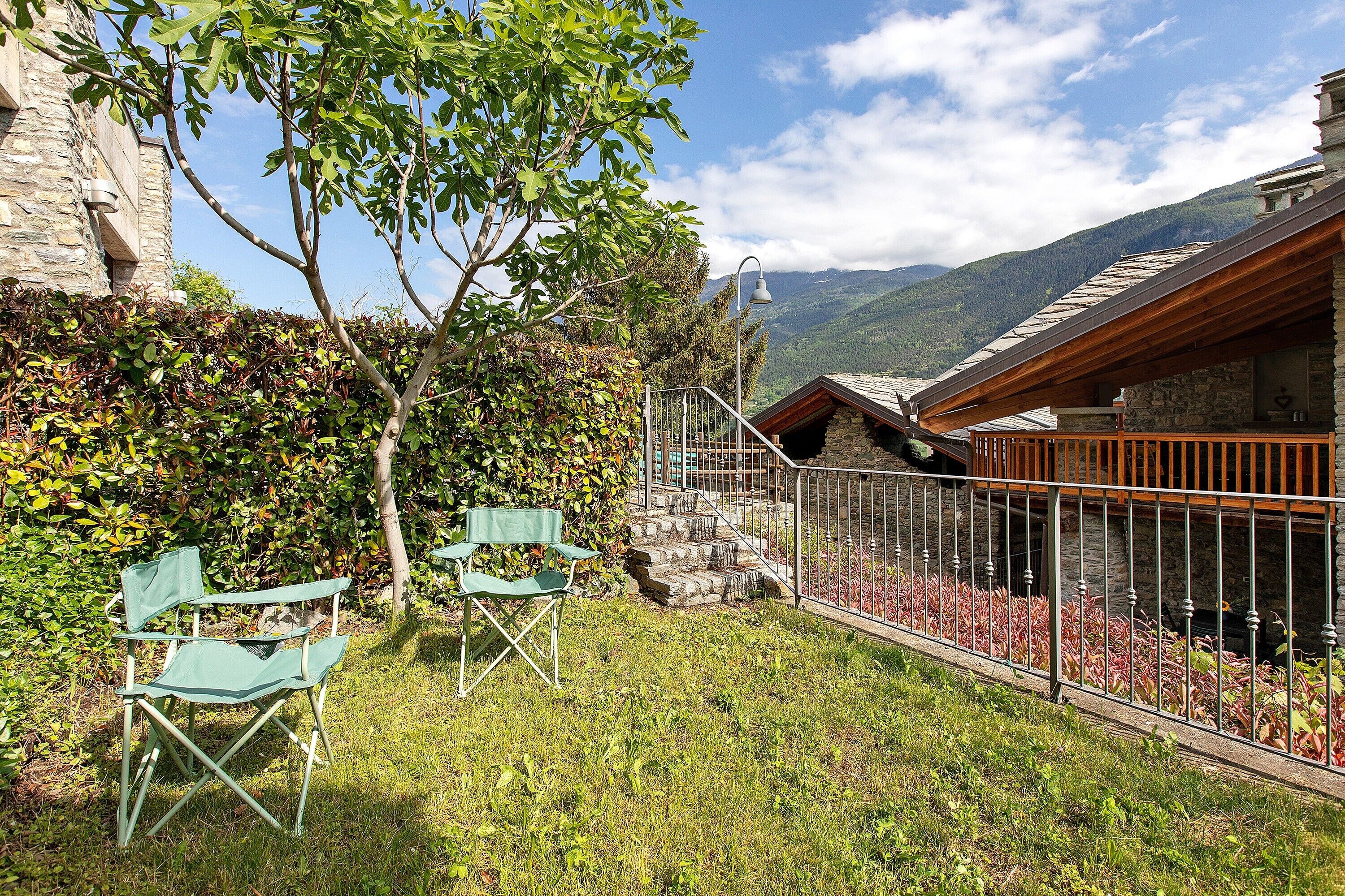 Un bâtiment en pierre doté d’un balcon en bois, un petit jardin avec de la pelouse et un arbre, et une vue sur les montagnes en arrière-plan.