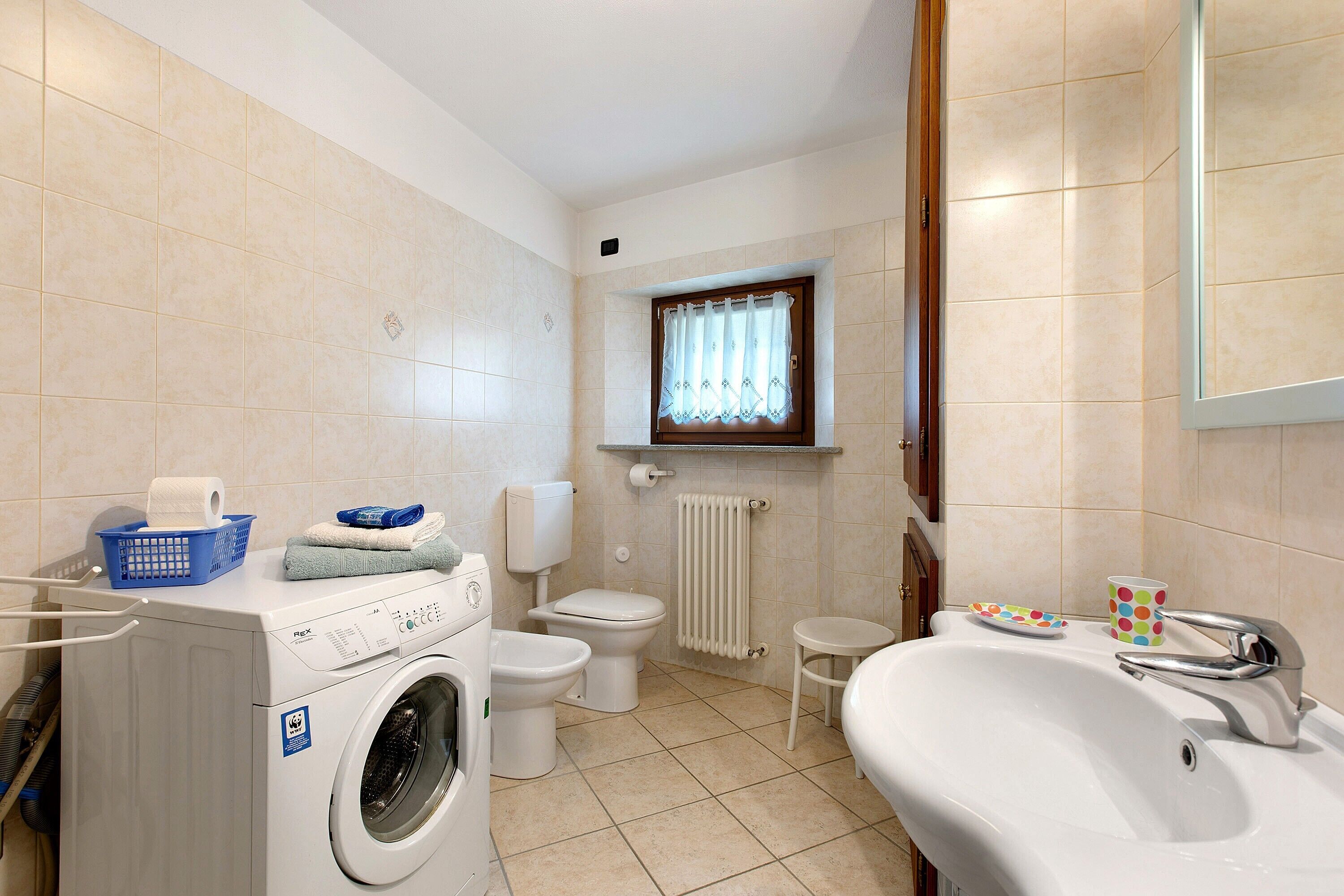 A bathroom with a washing machine, toilet, sink, and a window with a curtain.